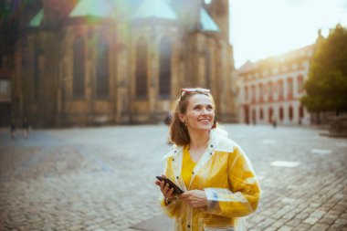 Prag 'da sarı bluz ve yağmurluk giyen gülümseyen genç bir kadın yürüyüş turu yapıyor, akıllı telefon kullanıyor ve yürüyor..