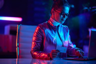 Neon metaevren fütüristik konsepti. Modern ofiste dizüstü bilgisayar kullanan, gözlüklü, modaya uygun bir kadın..