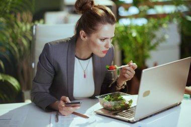 Sürdürülebilir iş yeri. Modern yeşil ofisteki gri takım elbiseli, modern orta yaşlı muhasebeci kadın. Laptop yiyor ve akıllı telefon kullanıyor..