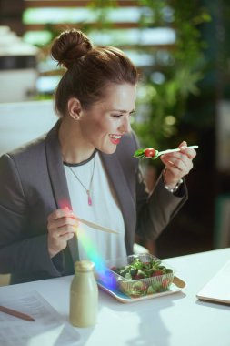 Sürdürülebilir iş yeri. Gülümseyen modern 40 yaşındaki muhasebeci kadın gri takım elbiseli, modern yeşil ofiste salata yiyor..