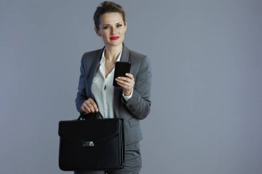Gri arka planda izole edilmiş akıllı telefon ve evrak çantasıyla gri takım elbiseli, güler yüzlü orta yaşlı kadın işçi..