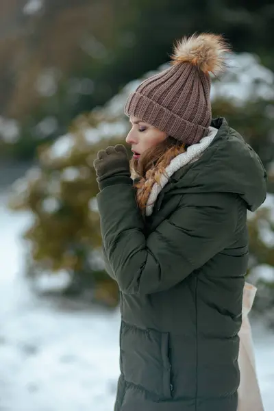 unhappy modern woman in green coat and brown hat outdoors in the city park in winter with mittens and beanie hat coughing.