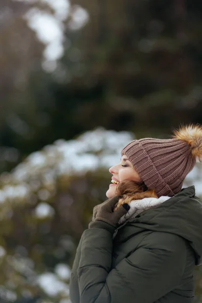 smiling modern woman in green coat and brown hat outdoors in the city park in winter with mittens and beanie hat.