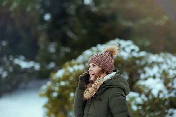 smiling modern woman in green coat and brown hat outdoors in the city park in winter with mittens and beanie hat near snowy branches talking on a smartphone.