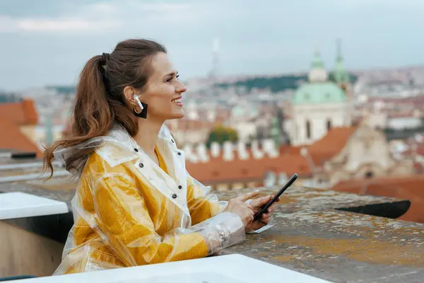 happy trendy solo traveller woman in yellow blouse and raincoat in Prague Czech Republic with headphones using smartphone against city panorama.
