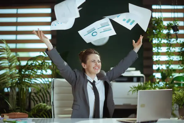 New job. smiling modern middle aged woman worker in modern green office in grey business suit with laptop throwing documents.