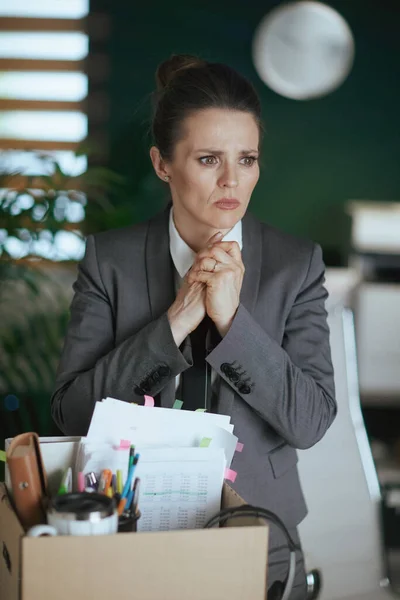 New job. sad modern 40 years old woman employee in modern green office in grey business suit with personal belongings in cardboard box.