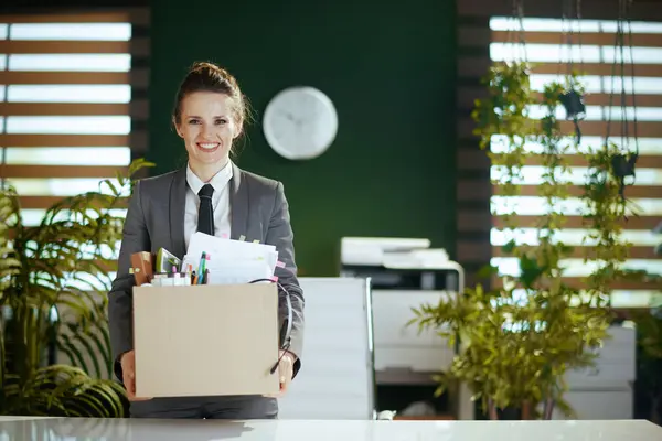 New job. happy modern female worker in modern green office in grey business suit with personal belongings in cardboard box.