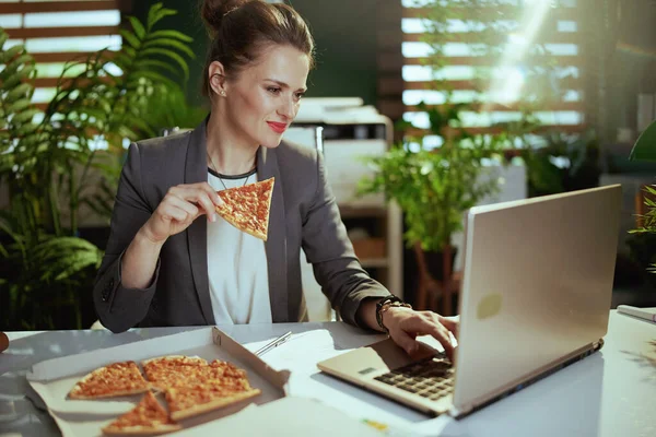 Sustainable workplace. modern small business owner woman in a grey business suit in modern green office with pizza and laptop.