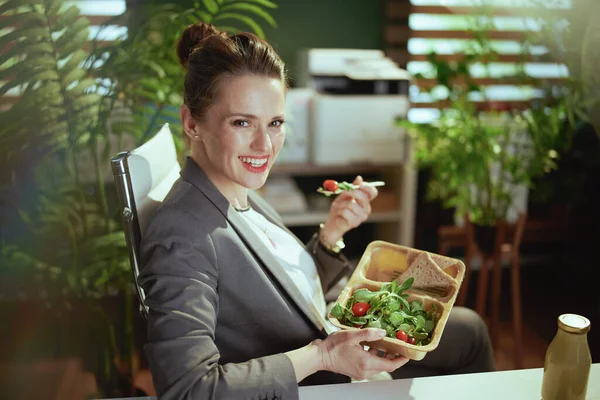 Sustainable workplace. happy modern woman worker in a grey business suit in modern green office eating salad.
