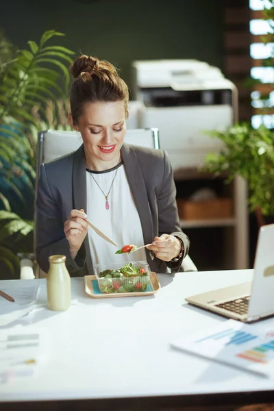 Sustainable workplace. happy modern middle aged woman worker in a grey business suit in modern green office with laptop eating salad.