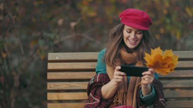 Merhaba sonbahar. Düşünceli, 40 yaşında, kırmızı şapkalı, güz yaprakları ve atkısı olan şehir parkında bankta otururken akıllı telefon kullanan bir kadın..