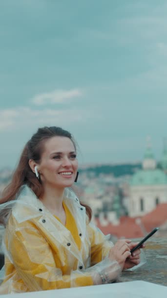 笑顔の若い旅行女性 黄色いブラウスとレインコート プラハチェコ共和国 都市パノラマに対するスマートフォンとヘッドフォン — ストック動画