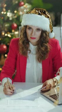 Noel zamanı. Gülümseyen orta yaşlı, Noel Baba şapkalı, kırmızı ceketli, modern yeşil ofiste Noel ağacıyla çalışan küçük işletme sahibi kadının portresi..