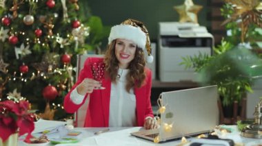 Noel zamanı. Noel Baba şapkalı, kırmızı ceketli, dizüstü bilgisayarı ve şarap bardağıyla mutlu, şık, küçük işletme sahibi bir kadın Noel ağacıyla modern yeşil ofiste web kamerası toplantısı yapıyor..