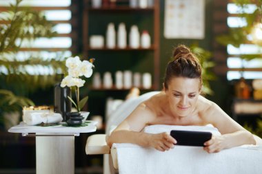 Sağlık hizmeti zamanı. Modern orta yaşlı kadın spa salonunda akıllı telefon kullanıyor ve masaj masasında yatıyor..