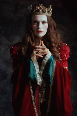 Kırmızı elbiseli, beyaz makyajlı ve tacı koyu gri arka planda olan ortaçağ kraliçesi..