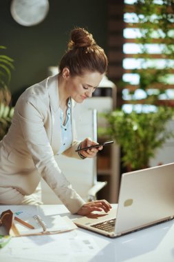 Modern yeşil ofisteki hafif takım elbiseli, akıllı telefon uygulaması kullanarak gülümseyen modern, küçük işletme sahibi kadın..