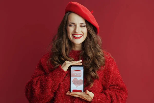 Happy Valentine. happy elegant female with long wavy hair in red sweater and beret , smartphone and Valentine app.