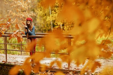 Merhaba sonbahar. Kot pantolonlu, kırmızı şapkalı, sonbahar yapraklı, atkılı, eldivenli ve çantalı zarif orta yaşlı kadının tam boy portresi..
