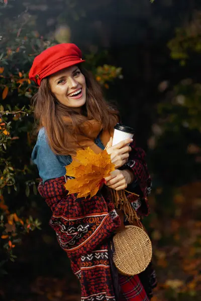 Hello autumn. smiling stylish middle aged woman in red hat with autumn leafs, scarf, bag and coffee in the city park.