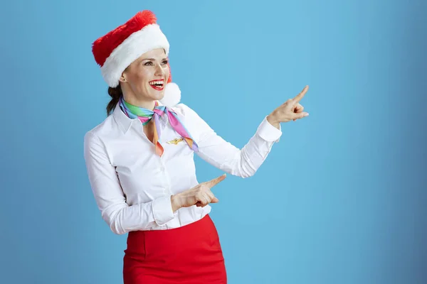 happy stylish stewardess woman against blue background in uniform with Santa hat pointing at something.