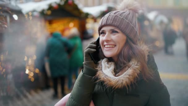在这个城市的冬季展会上 穿着绿色外套 头戴褐色帽子的现代女性用智能手机微笑着 — 图库视频影像