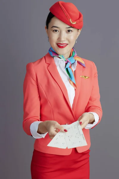 smiling modern air hostess asian woman in red skirt, jacket and hat uniform with flight tickets isolated on grey background.