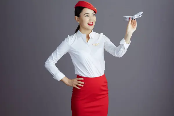 happy elegant flight attendant asian woman in red skirt and hat uniform with a little airplane isolated on gray.