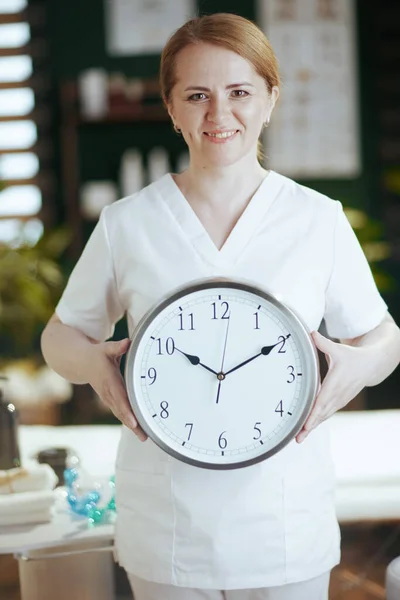 Healthcare time. happy female medical massage therapist in massage cabinet with clock.