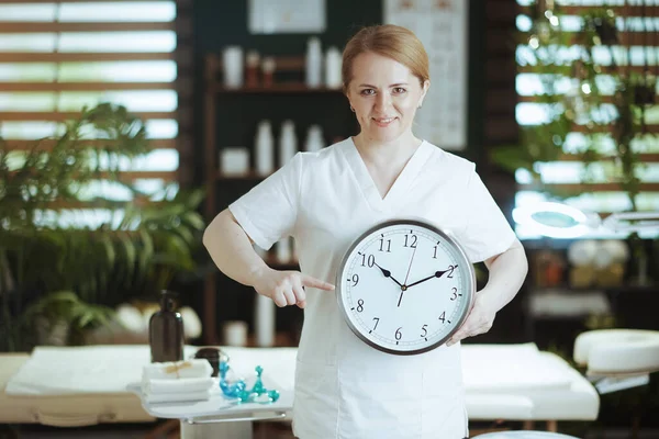 Healthcare time. smiling female medical massage therapist in massage cabinet with clock.