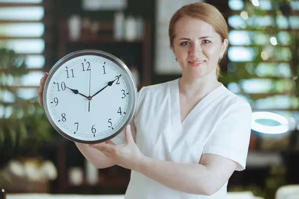 Healthcare time. Portrait of smiling massage therapist woman in massage cabinet with clock.