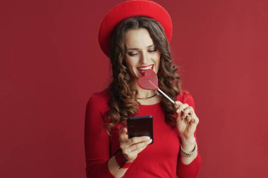 Mutlu Sevgililer. Kırmızı elbiseli, uzun dalgalı saçlı ve bereli mutlu modern bir kadın. Kırmızı arka planda kalp şeklinde şekerler ve akıllı telefon kullanarak kısa mesaj yolluyor..