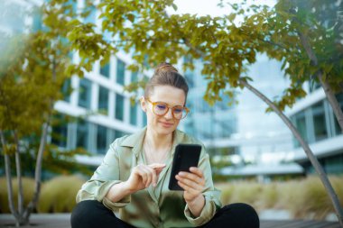 Yeşil bluzlu ve gözlüklü iş merkezinin yakınında akıllı telefon kullanan düşünceli modern kadın işçiler..