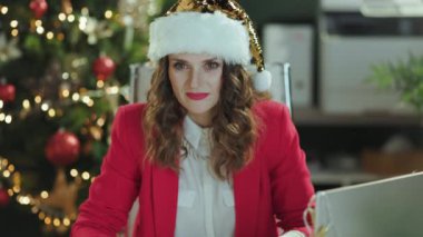 Noel zamanı. Gülümseme portresi kırmızı ceket içinde Noel Baba şapkası içinde modern yeşil ofiste Noel ağacı ile.