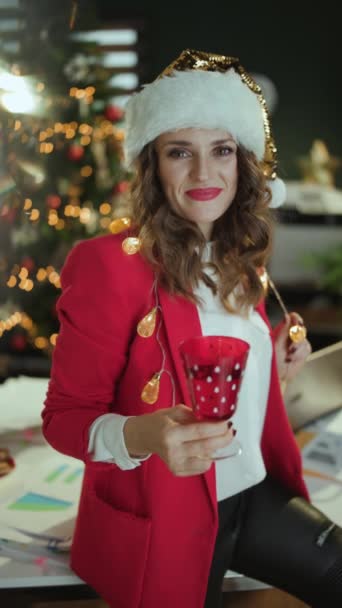 クリスマス タイム エレガントな 歳のビジネス女性 サンタ帽子と赤いジャケット クリスマスツリーと現代グリーンオフィスでワイングラスダンス — ストック動画