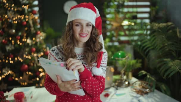 クリスマス タイム ハッピースタイリッシュな小さなビジネスオーナーの女性の肖像画 サンタの帽子と赤のクリスマスセーター 現代緑のオフィスのドキュメントとクリスマスツリー — ストック動画