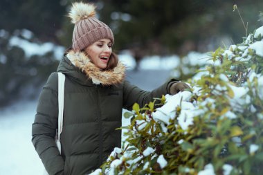 Gülümseyen orta yaşlı, yeşil ceketli ve kahverengi şapkalı bir kadın kışın şehir parkında eldivenleri ve karlı dalların yanında şapka takıyor..