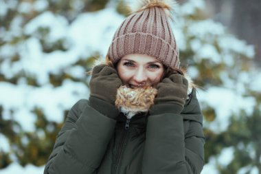 Gülümseyen 40 yaşındaki yeşil paltolu ve kahverengi şapkalı kadın kışın şehir parkında eldivenleri ve karlı dalların yanında şapka takıyor..