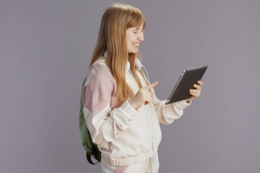 Sırt çantası ve kulaklıklarıyla gri üzerine izole edilmiş tablet kullanan bej eşofmanlı modern okul kızı gülümsüyor..