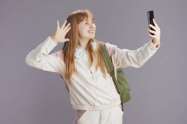 Sırt çantası ve kulaklıklı, gri el sallayarak izole edilmiş akıllı telefonuyla selfie çeken bej eşofmanlı mutlu modern bir genç kadın..