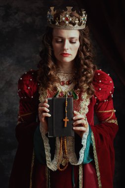 Kırmızı elbiseli ortaçağ kraliçesi koyu gri arka planda kitap, tespih ve taçla.