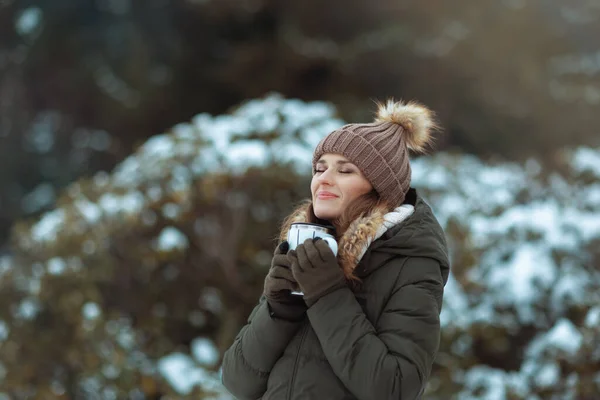 快乐的现代女性 冬天穿着绿色外套 戴着棕色帽子 头戴手套 戴着热可可 头戴便帽 站在白雪公主的枝头附近 — 图库照片