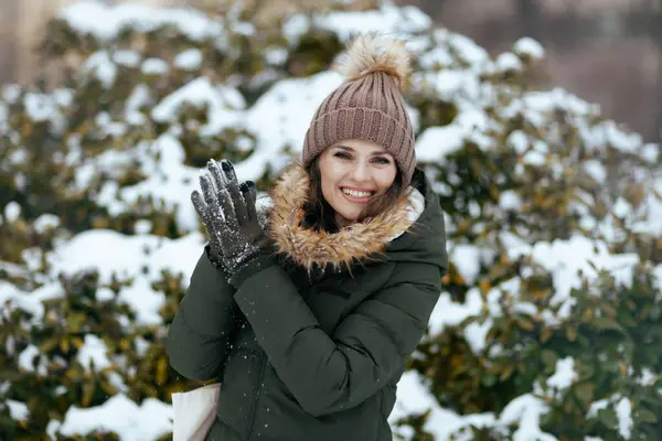 happy modern woman in green coat and brown hat outdoors in the city park in winter with mittens and beanie hat near snowy branches.