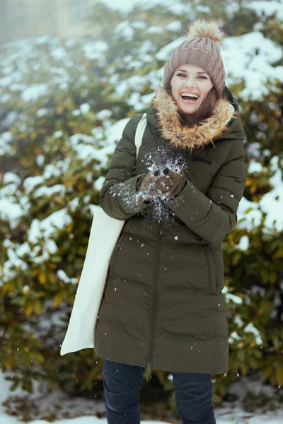 smiling modern woman in green coat and brown hat outdoors in the city park in winter with mittens and beanie hat playing with snow near snowy branches.