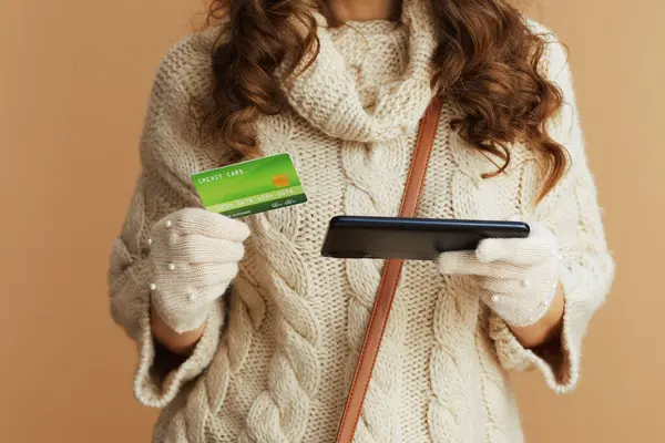 こんにちは ベージュセーターの女性のクローズアップ スマートフォンとクレジットカードブラウジングオンラインショップでベージュの背景にミトンと帽子 — ストック写真