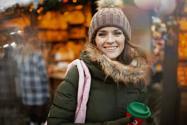 happy modern woman in green coat and brown hat at the winter fair in the city with Christmas beverage.