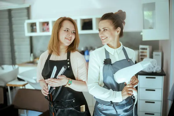 smiling two 40 years old women hairdressers in aprons in modern beauty salon with hair straightener and hair dryer.