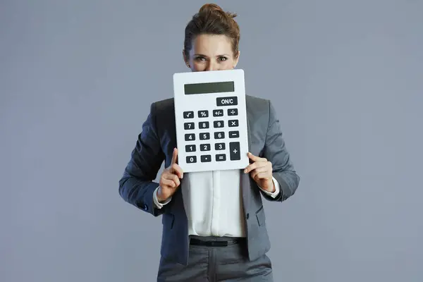 elegant middle aged woman worker in grey suit with calculator against gray background.