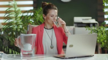 Sürdürülebilir iş yeri. Gülümseyen modern, küçük işletme sahibi bir kadının portresi. Kırmızı ceketli, su filtresi ve dizüstü bilgisayarlı..